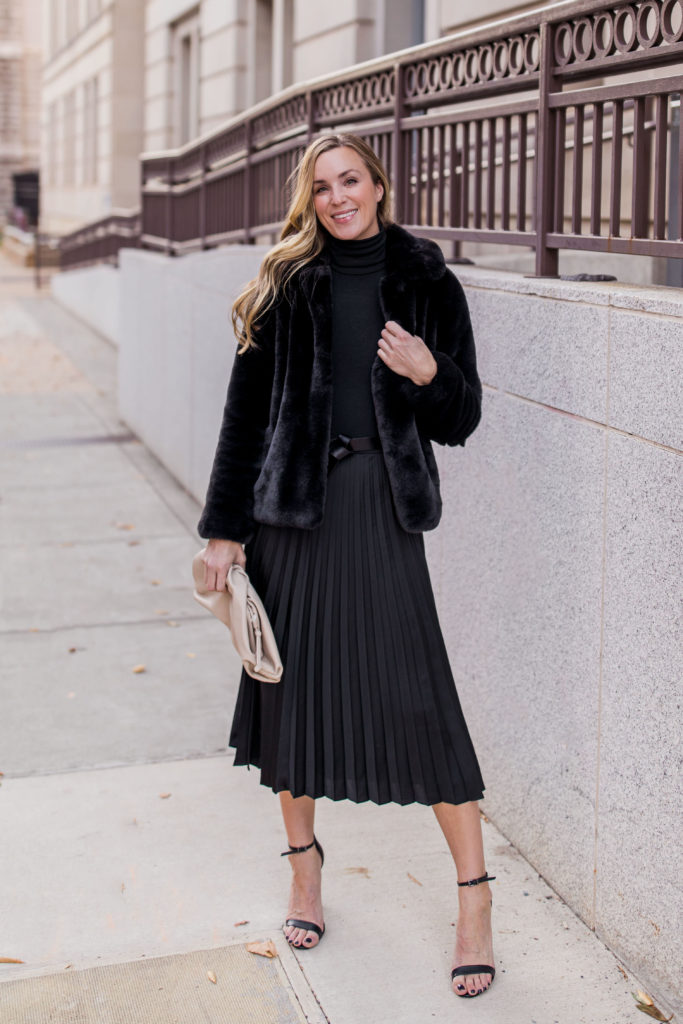 woman wearing faux fur jacket with black pleated midi skirt and black turtleneck